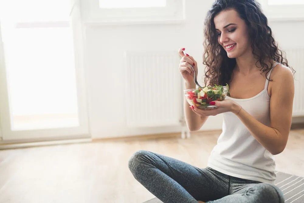 A beautiful woman with a curly hair eating salad on a gym sitting on a wooden floor with a window view on the background